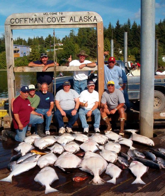 Group under the Welcome to Coffman Cove Alaska sign with a full spread of halibut